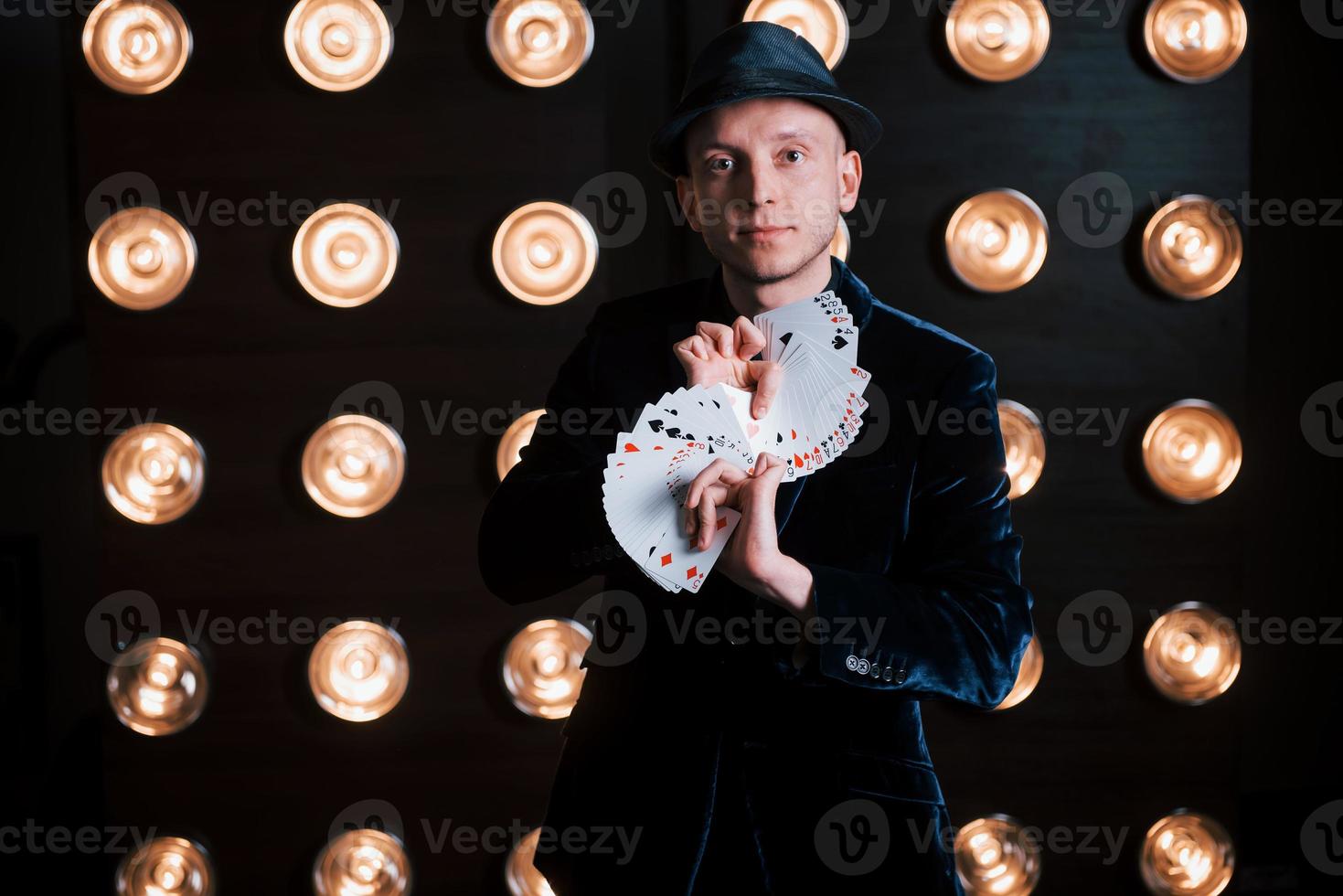 concepción del ilusionista. mago en traje negro y con cartas de juego de pie en la habitación con iluminación especial en el backstage foto