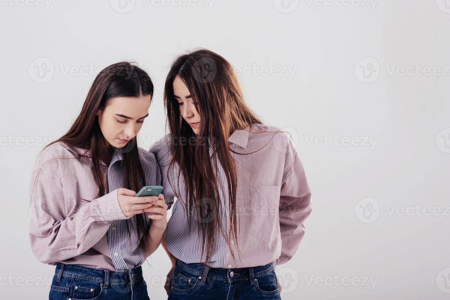 Enters something in the smartphone. Two sisters twins standing and posing in the studio with white background photo