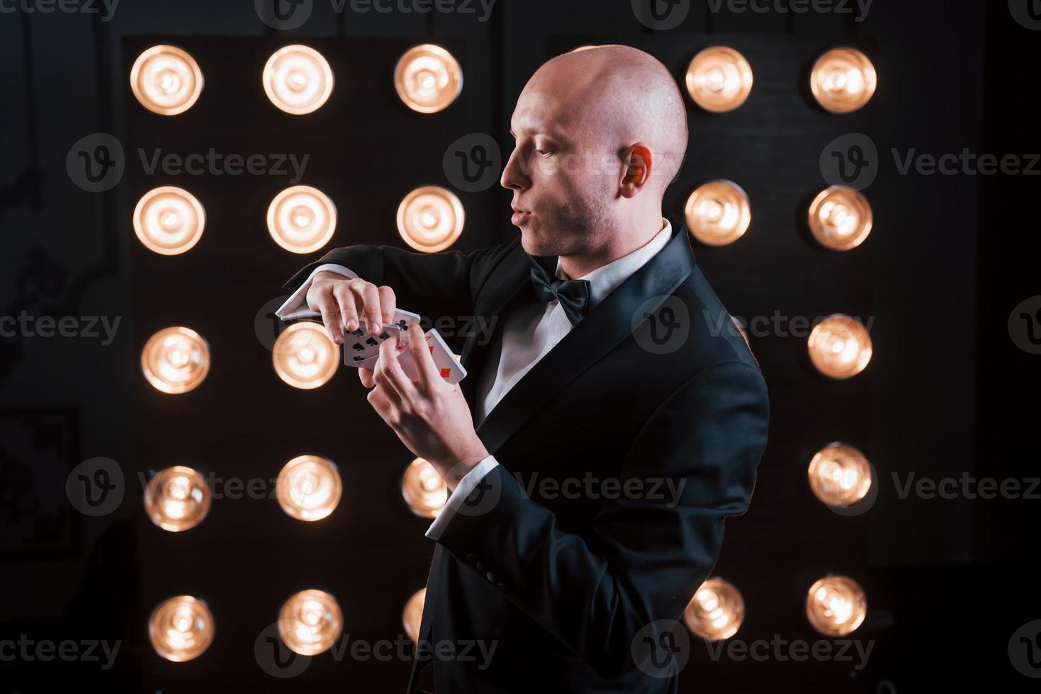 Concentrating at tricky move. Magician in black suit and with playing cards standing in the room with special lighting at backstage photo