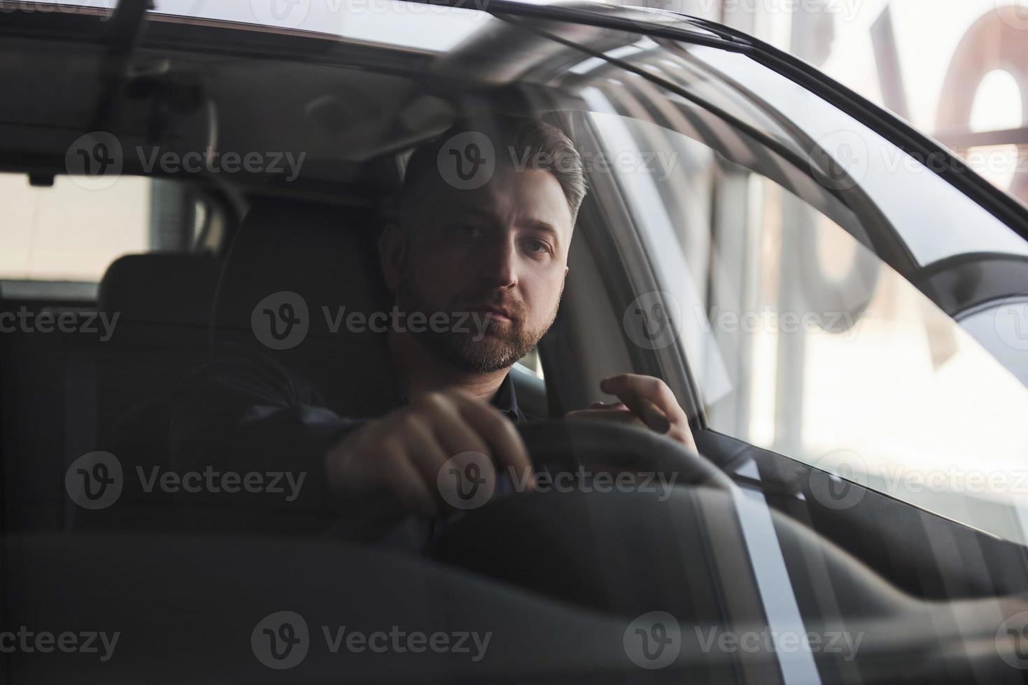 relajándose con cigarro en mano. vista frontal de un joven hombre de negocios barbudo sentado en su auto negro de lujo foto