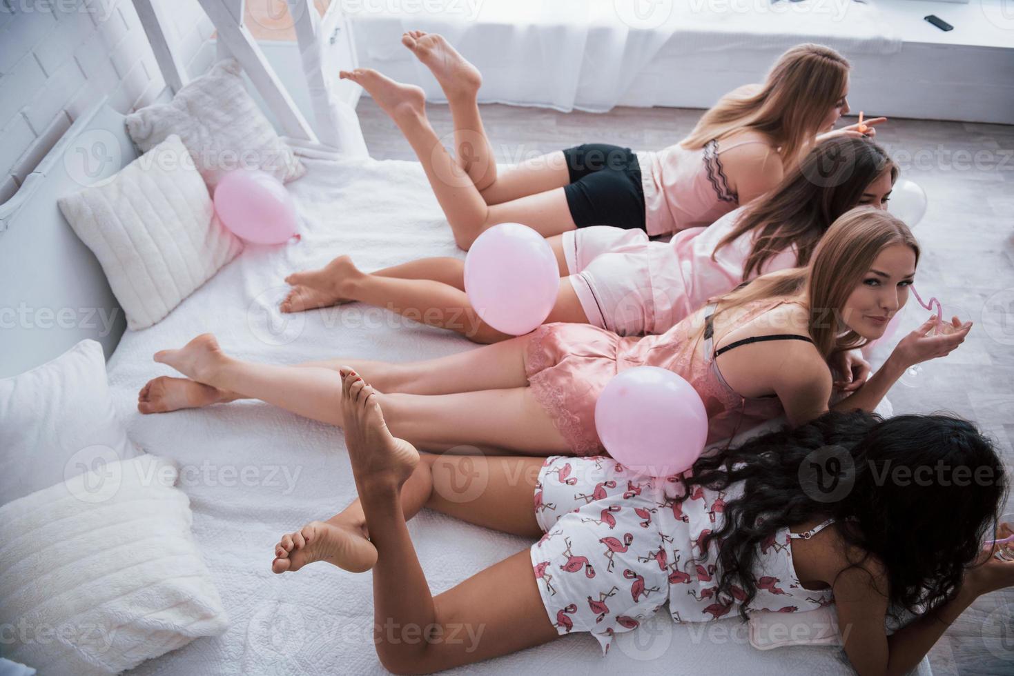 sosteniendo una bebida. las chicas jóvenes acostadas en una cama blanca de lujo tienen una celebración. vista lateral y superior foto