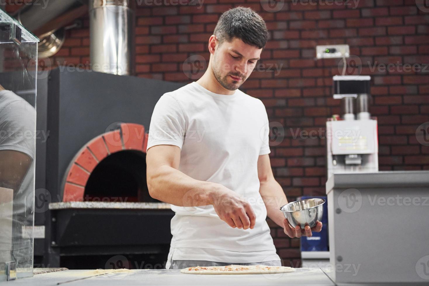 Giving some solt. Baker in white shirt throws spices to make delicious pizza for an order in restaurant photo