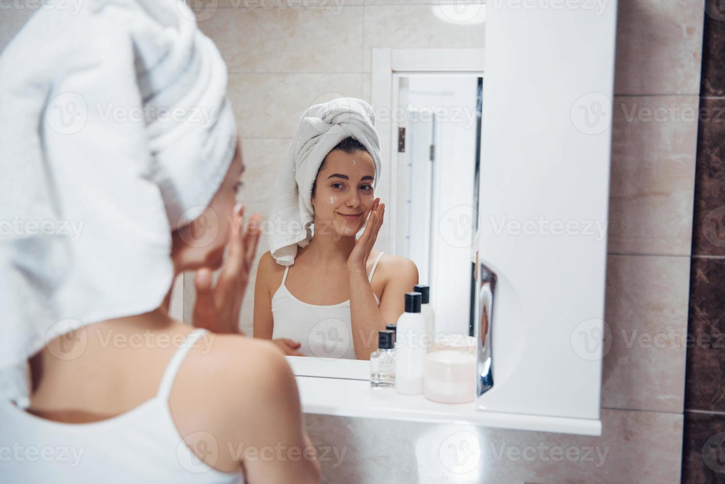 poner crema en la cara. en el baño. mujer con toalla blanca en la cabeza tiene cuidado de sí misma foto