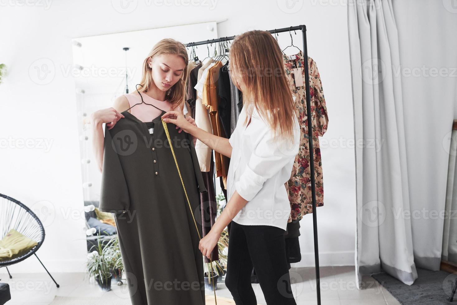 Measures height of black wear. Two girls working on the order in sewing shop with many clothes at daytime photo
