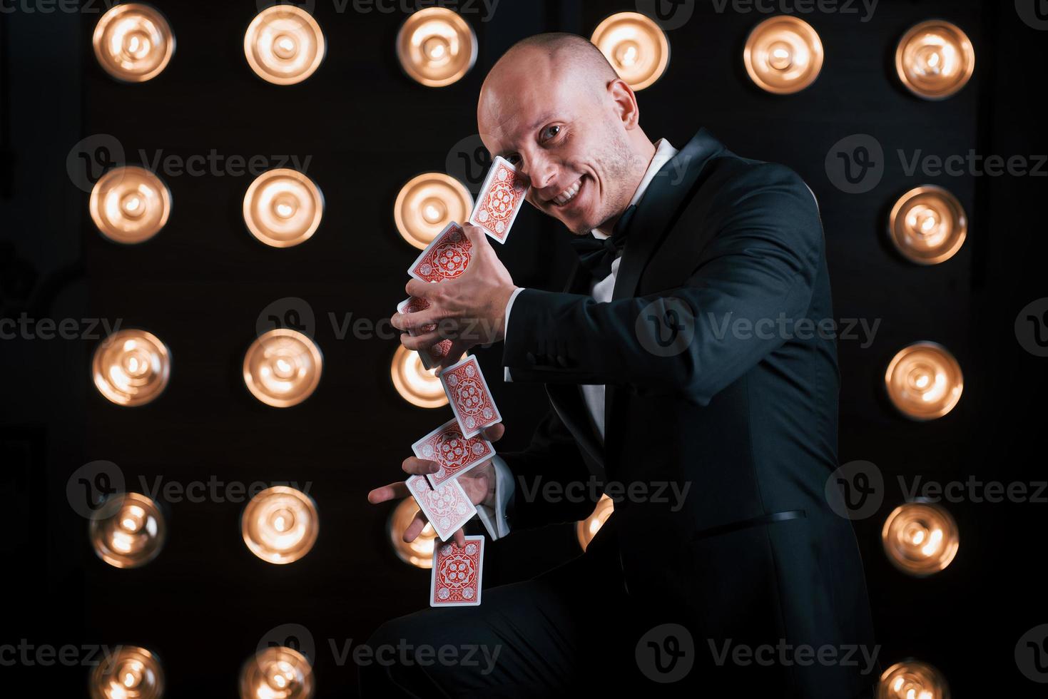 sonriendo durante el proceso. mago en traje negro y con cartas de juego de pie en la habitación con iluminación especial en el backstage foto