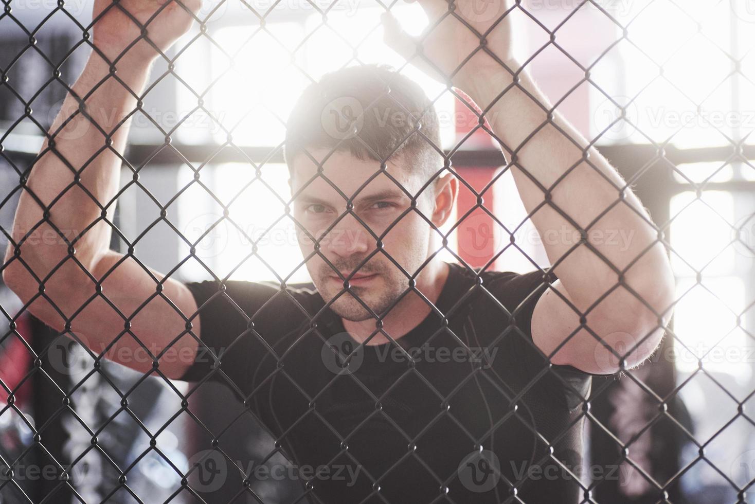 Close up view. Sportive guy in black shirt stands behind the fence in the room with daylight behind photo