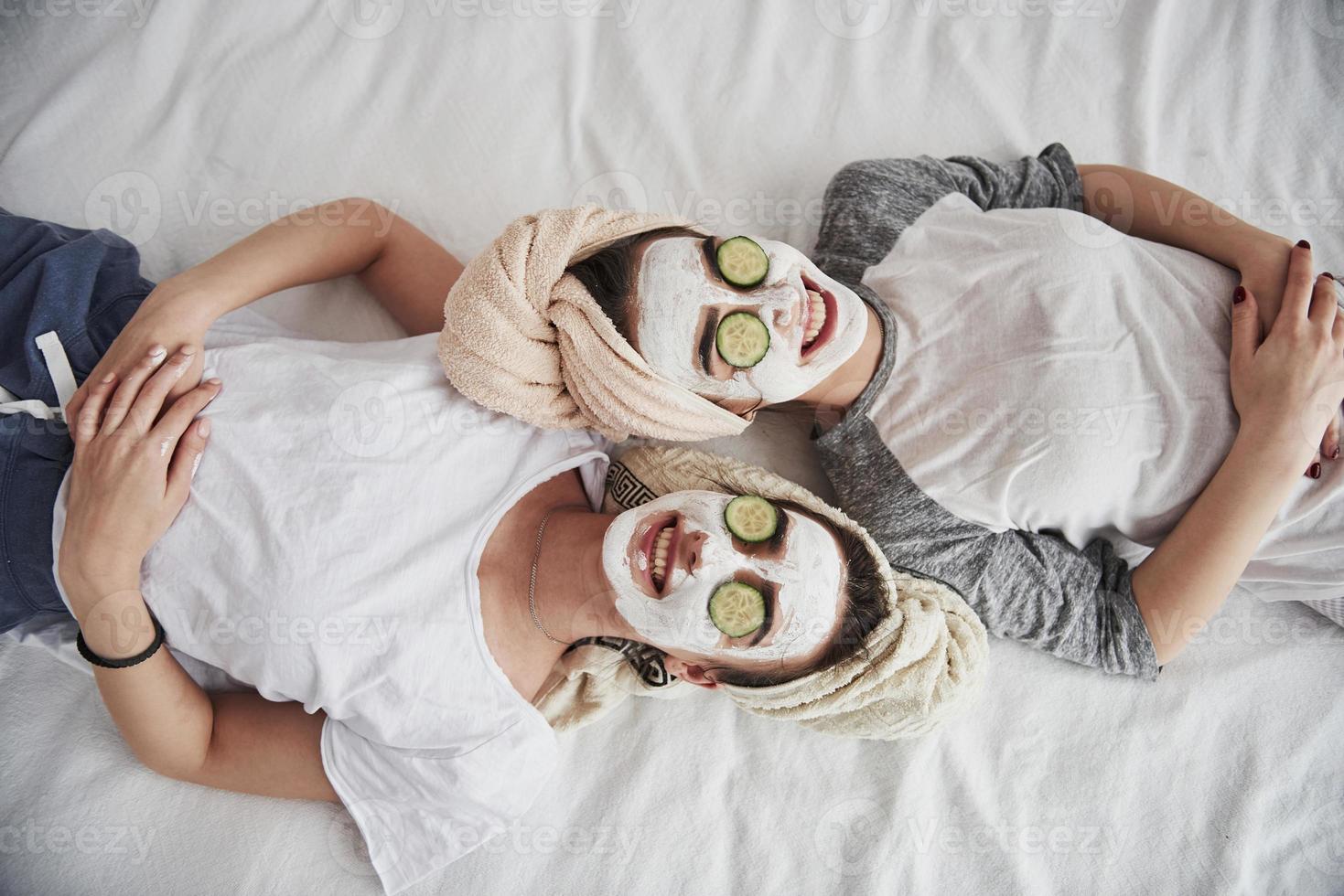Everyone is smiling. Lying on the white bed. Top view. Conception of skin care by using white mask and cucumbers on the face photo