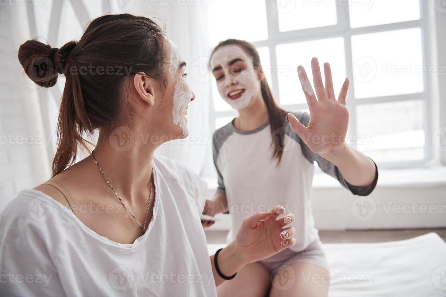 sonriendo durante el proceso. concepción del cuidado de la piel mediante el uso de máscara blanca en la cara. dos hermanas mujeres tienen fin de semana en el dormitorio foto