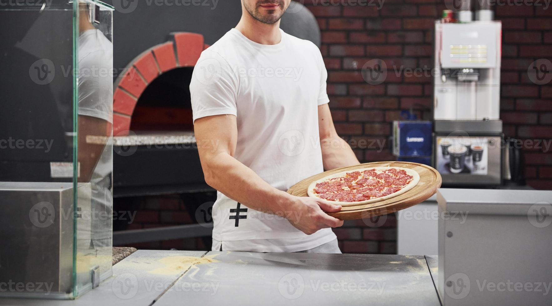 Holding in strong hands. Baker in white shirt with pizza that ready for put in the oven to cook photo