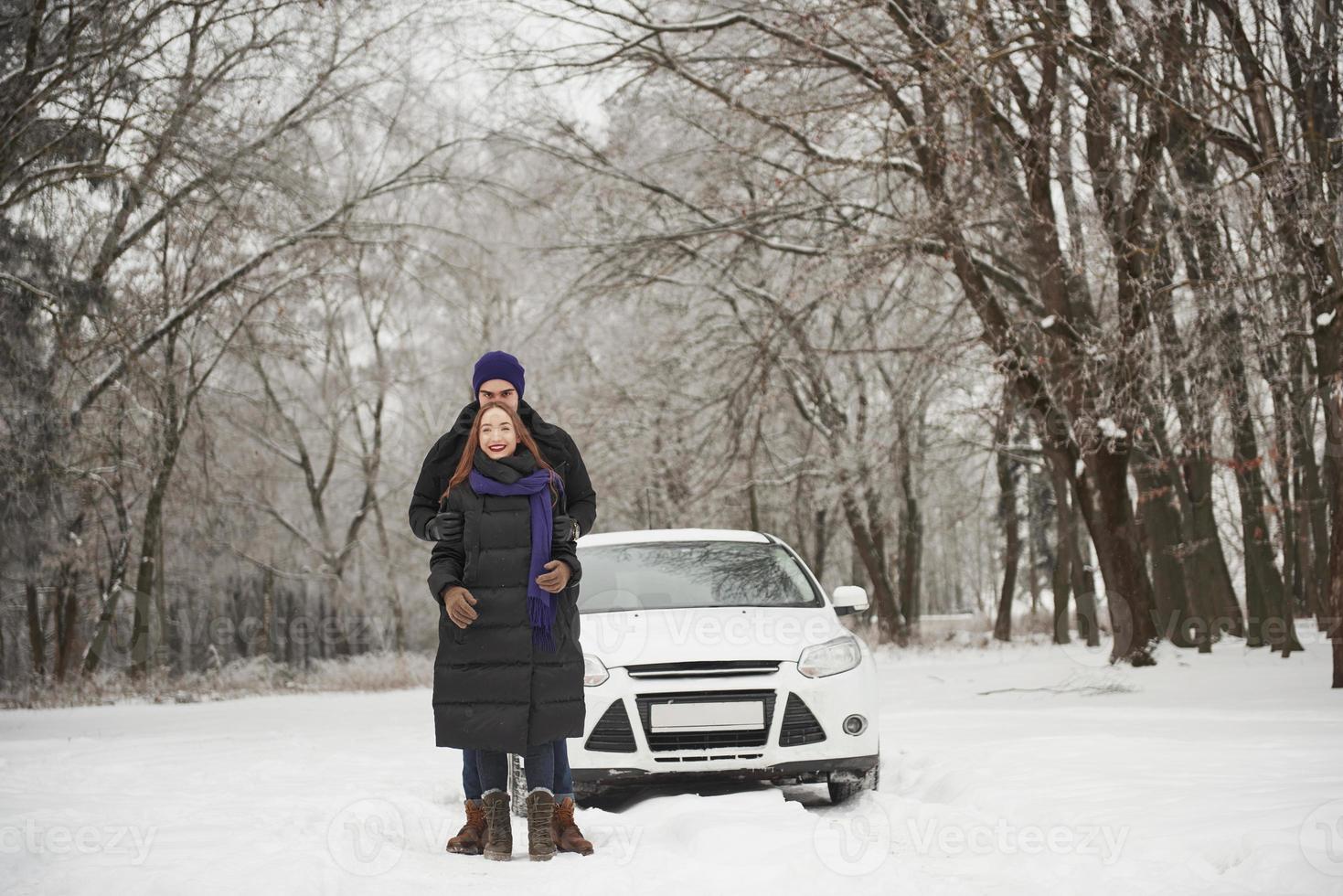 Gorgeous young couple stands in front of their white automobile in snowy forest photo