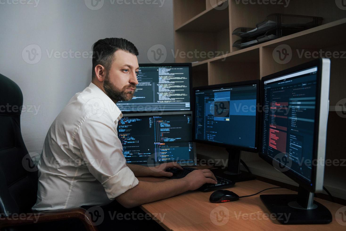 códigos de programa está en todas partes. hombre barbudo con camisa blanca trabaja en la oficina con múltiples pantallas de computadora en gráficos de índice foto