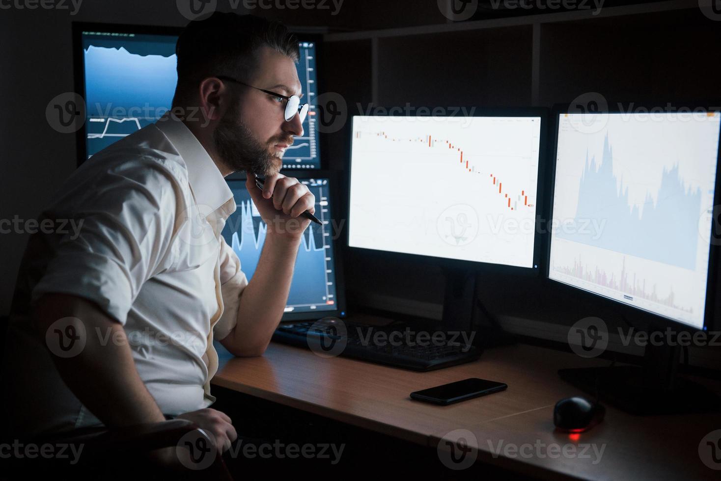 Thoughtful look. Bearded man in white shirt works in the office with multiple computer screens in index charts photo