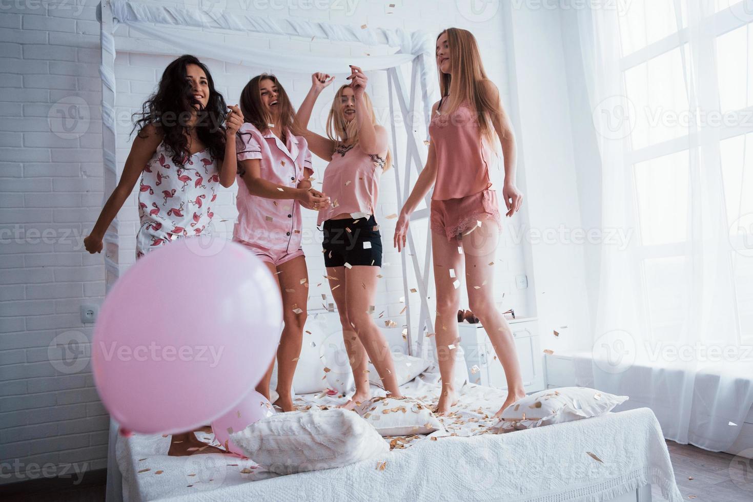 Pink balloon on the left side. Confetti in the air. Young girls have fun on the white bed in nice room photo