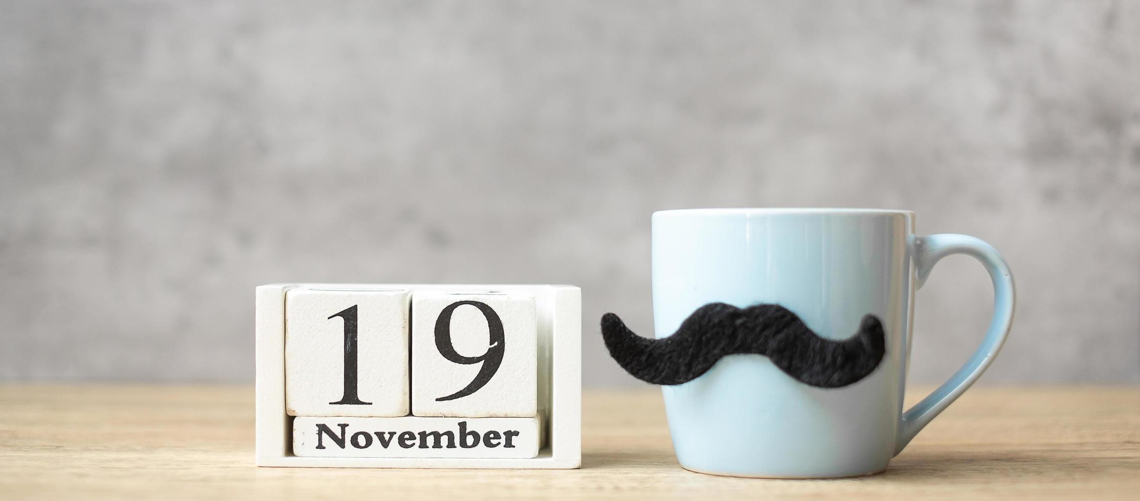 día internacional del hombre con calendario del 19 de noviembre, taza de café azul o taza de té y decoración de bigote negro en la mesa. feliz día del padre y concepto de celebración foto