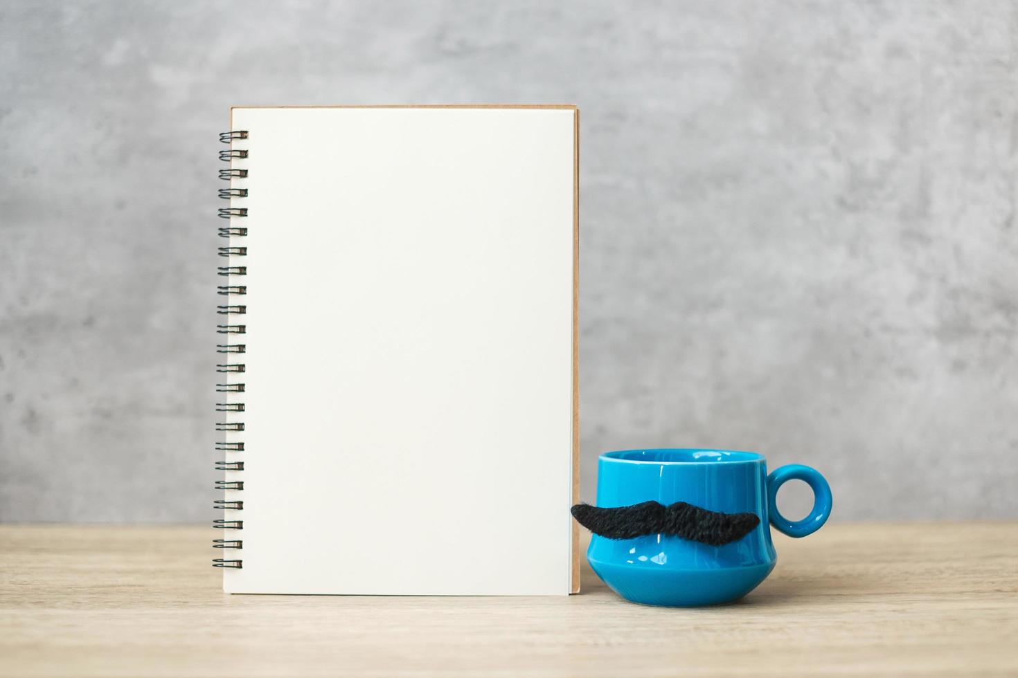 Blue coffee cup or tea mug with Black mustache decor and empty paper notepad or calendar on table. Blank copy space for text. Blue November, Happy Father day and International men day concept photo