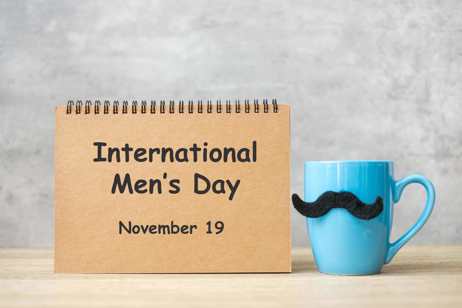 día internacional del hombre con bloc de notas de papel, taza de café azul o taza de té y decoración de bigote negro en la mesa. feliz día del padre y concepto de celebración foto