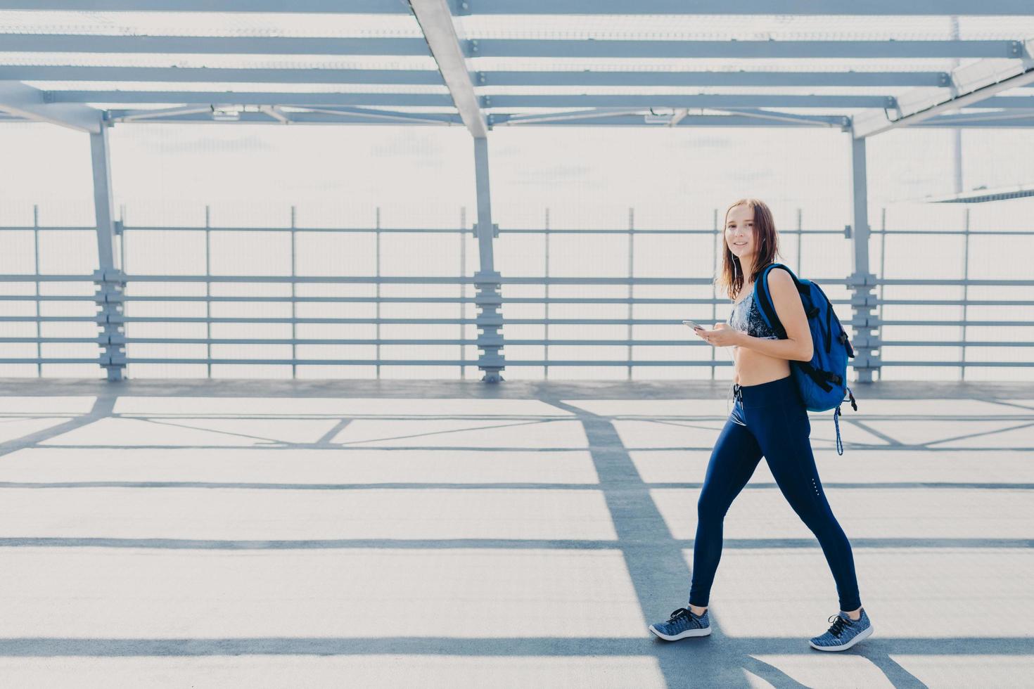 Outdoor shot of beautiful woman wth slim body, dressed in casual top and leggings, carries bag, going in gym, uses modern smart phone for listening music, copy space aside for your advertisement photo