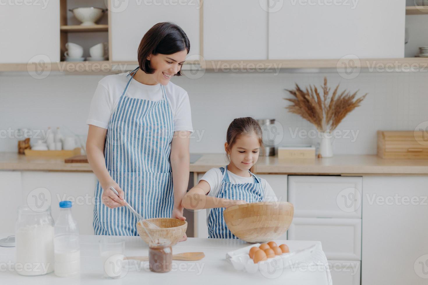 foto de la madre morena y el niño pequeño hacen masa para galletas, baten los ingredientes en un tazón, se visten con delantales, mamá se regocija de tener un pequeño ayudante en la cocina. cocinar juntos, familia y concepto de hogar