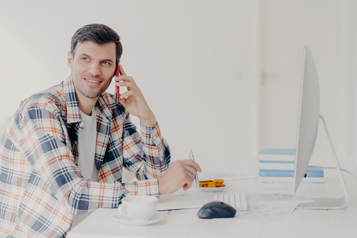 Horizontal shot of busy male freelancer has telephone conversation, dressed in checkered shirt, writes down information, sits at desktop with computer, solves financial problem, poses at workplace photo