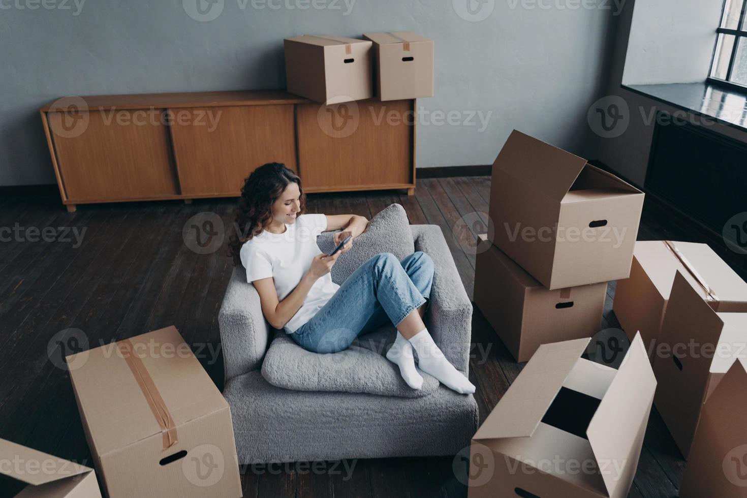 Girl having rest in new apartment and clicks phone. Independent spanish woman is moving. photo