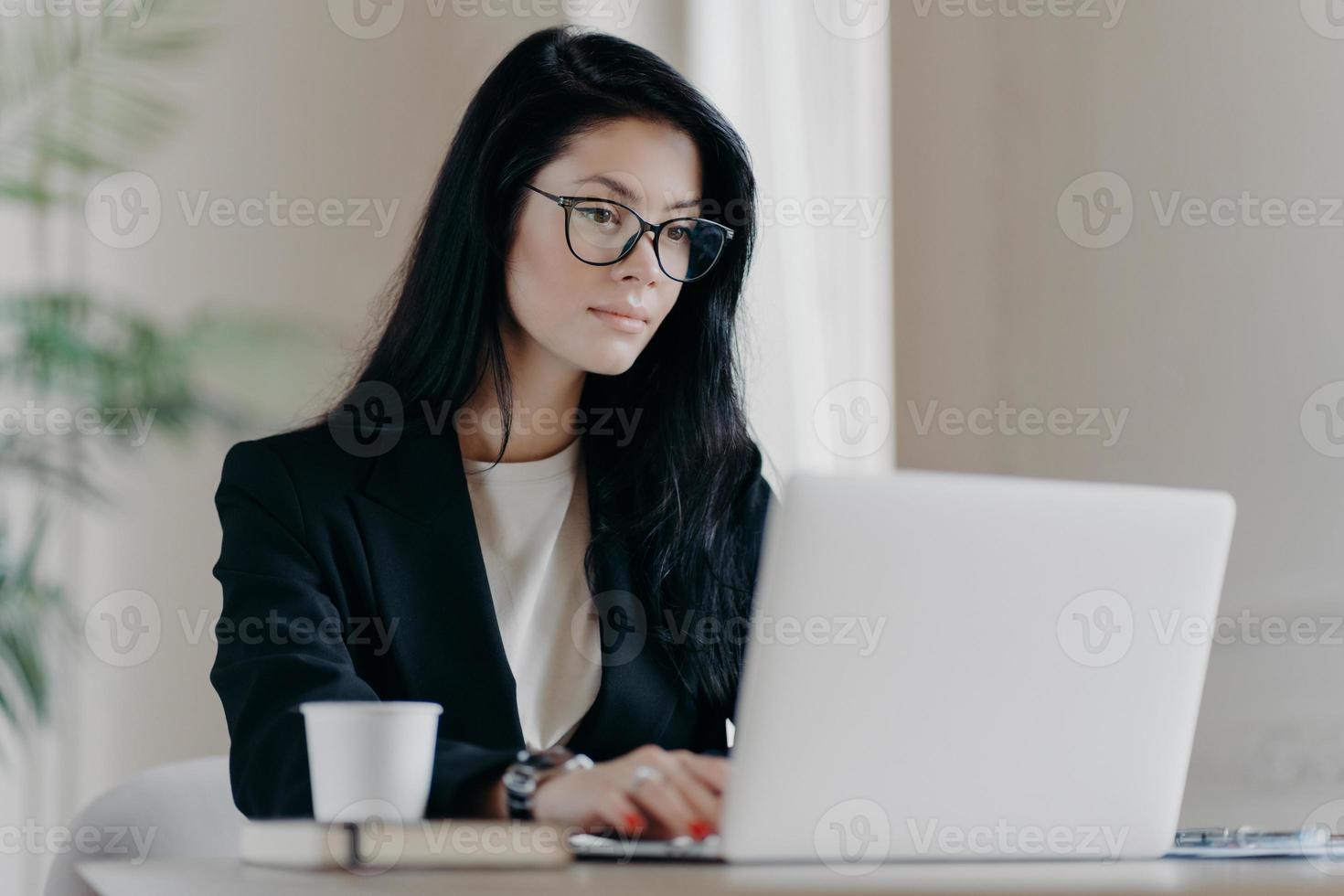 Concentrated brunette woman IT specialist in eyewear searches information using internet, keyboards on laptop computer, drinks coffee, has serious expression. Business and freelance concept. photo