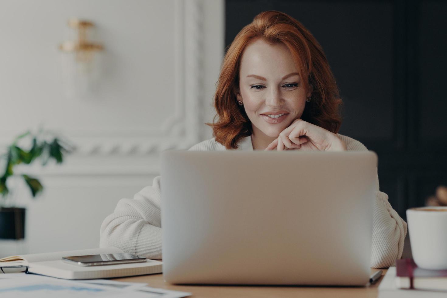 Experienced red haired businesswoman reads publication on modern laptop computer, concentrated at screen with pleasant smile, makes research for planning startup, checks mail box, works from home photo