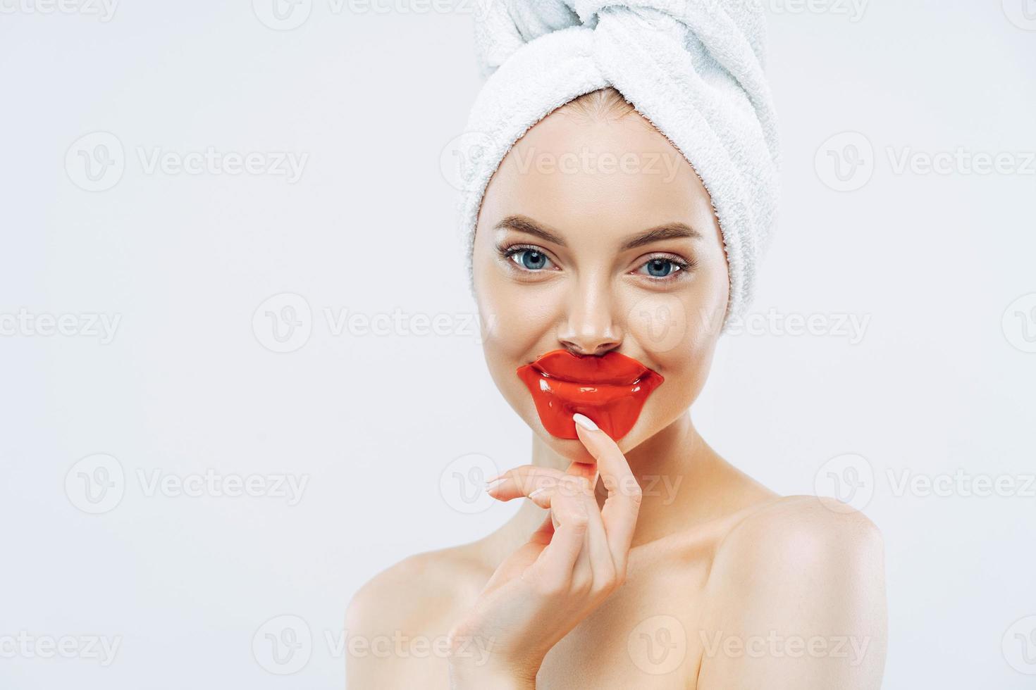 Pleasant looking healthy woman keeps patches on lips, wears towel on head, has clean pure skin after hygienic procedure, stands shirtless against white background, empty space for your advertisement photo