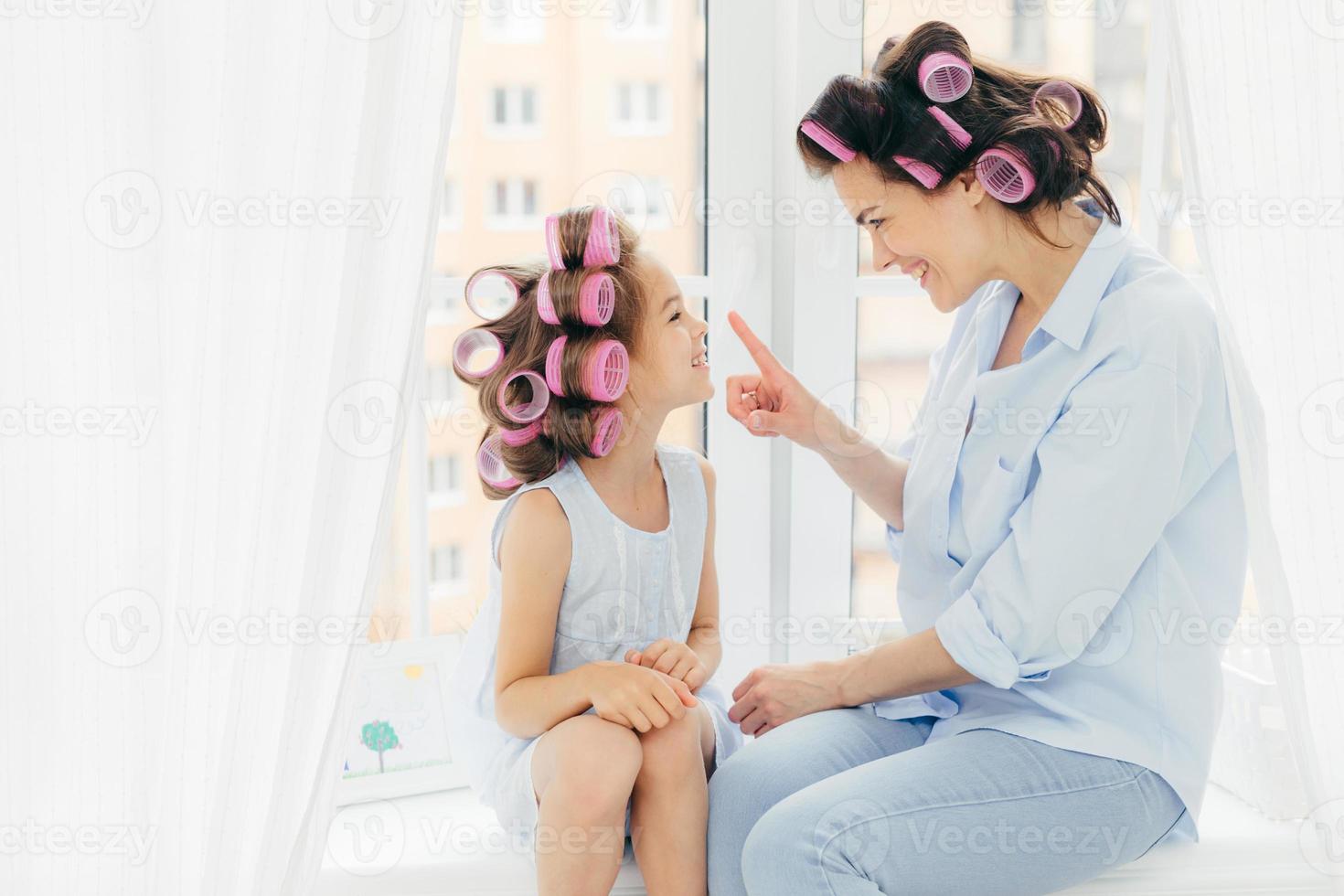 Cheerful two mother and daughter do hair nd have fun together, have curlers on head, sit on window sill. Affectionate mum touches nose of her small kid, prepare for party, care of hair and beauty photo
