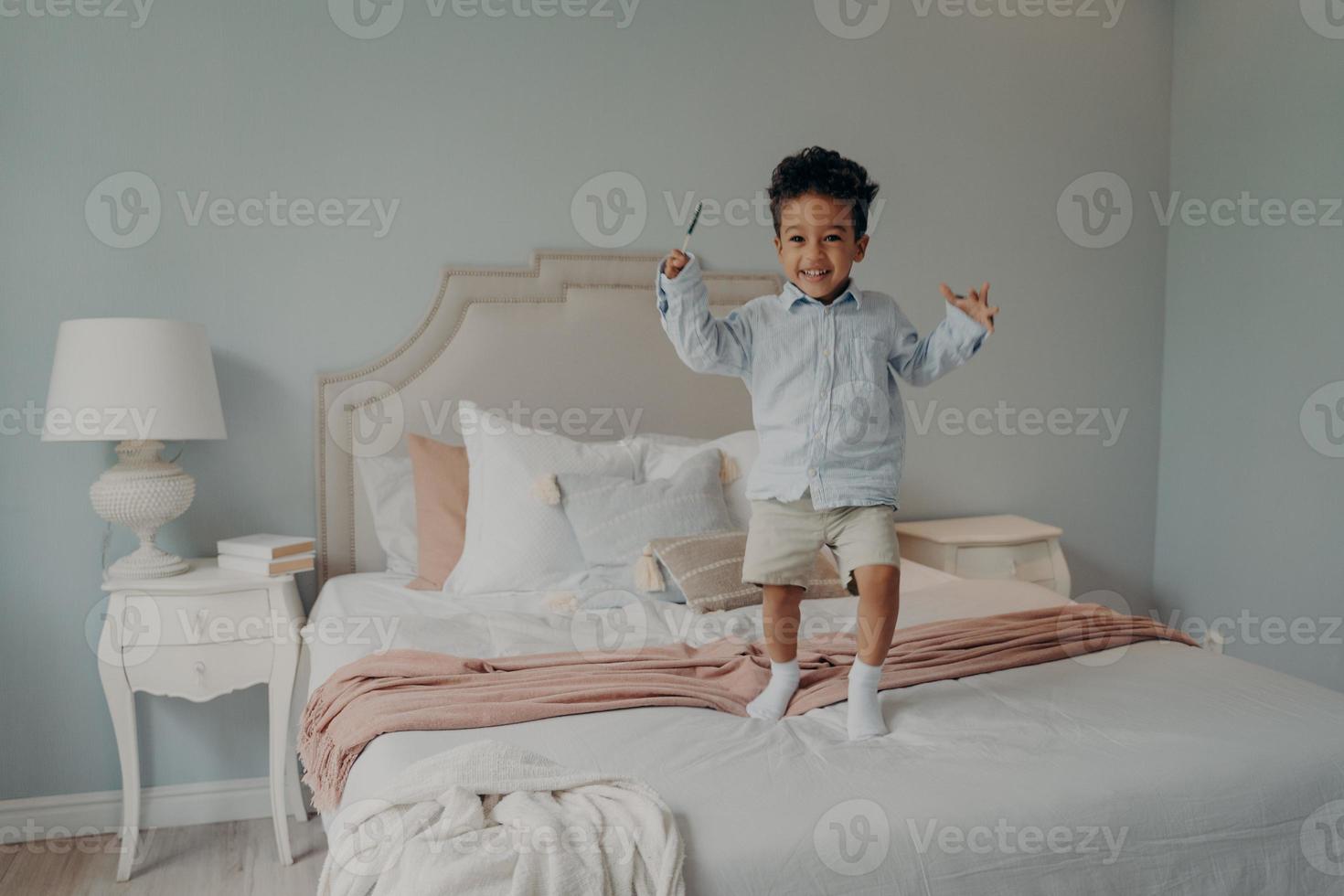 Joyful small afro american kid jumping on bed at home and smiling photo