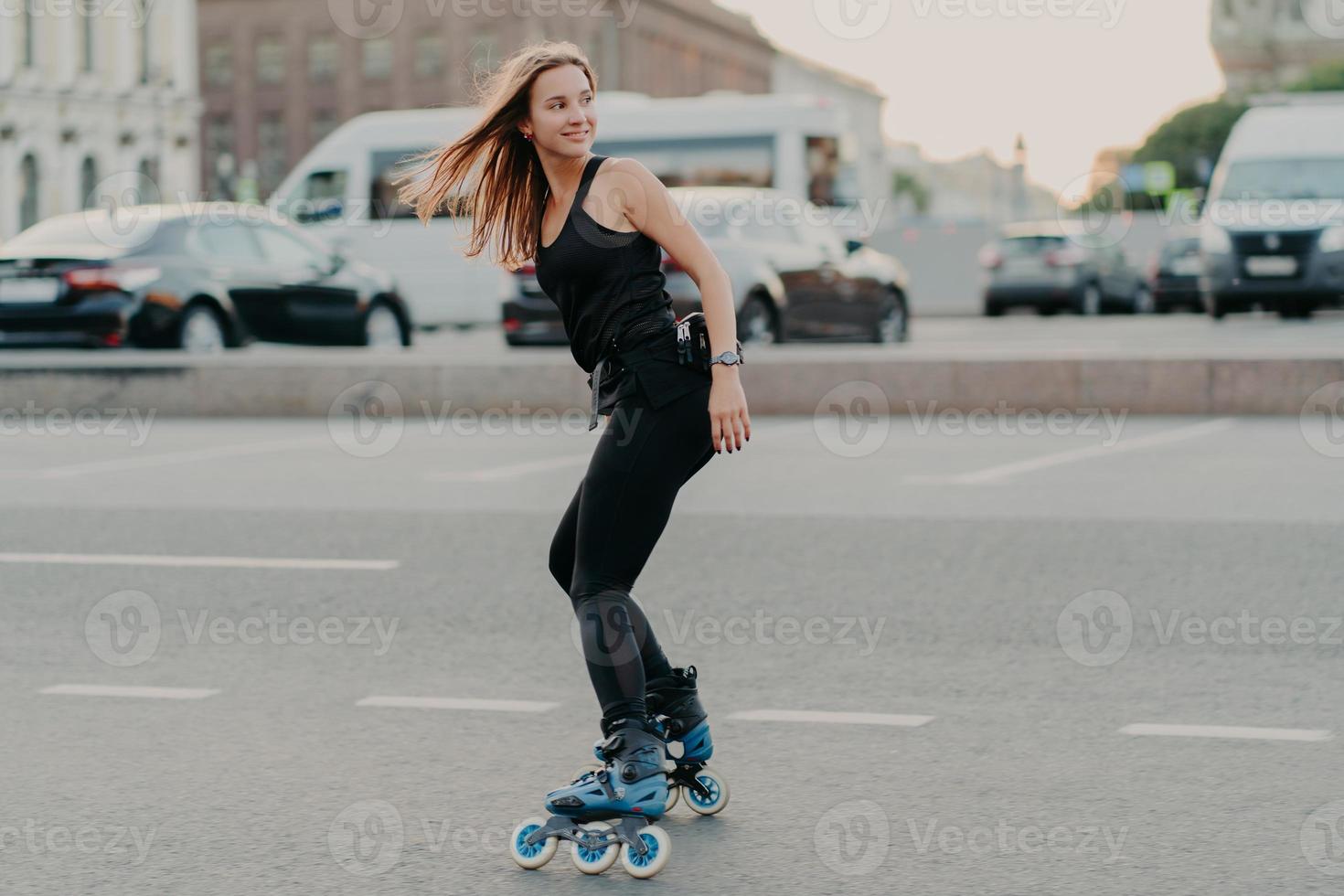 una foto lateral de una joven activa en patines en buena forma física a lo largo de las calles de la ciudad se ve feliz detrás, usa ropa deportiva negra y lleva un estilo de vida saludable. concepto de recreación y hobby