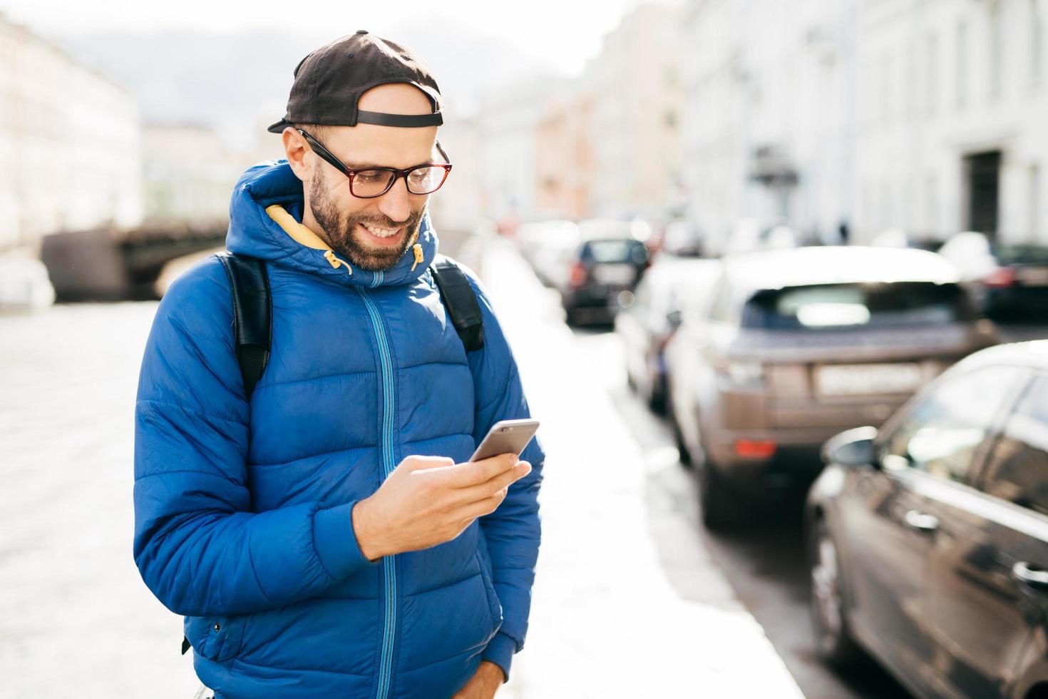 Young bearded hipster guy in blue anorak and cap holding smartphone answering call having happy expression isolated over big city background. Stylish man using cell phone while standing on street photo