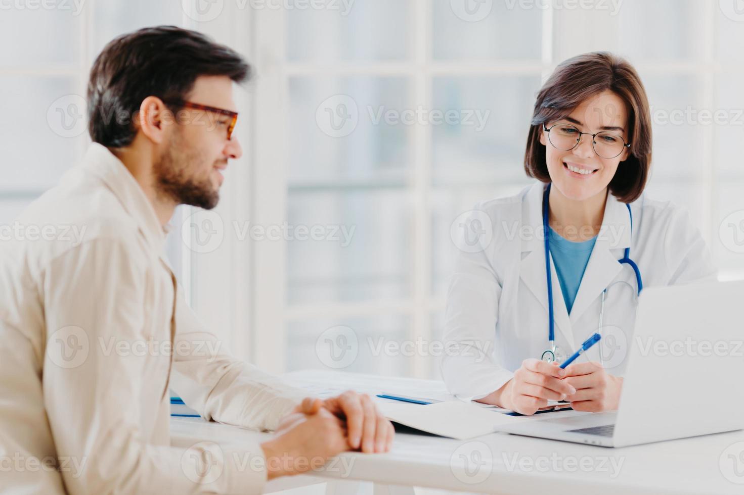 Horizontal shot of doctor and patient discuss something, talk about diagnosis, use laptop computer, pose in consulting room, check medical records. Sick man gets prescription from therapist. photo