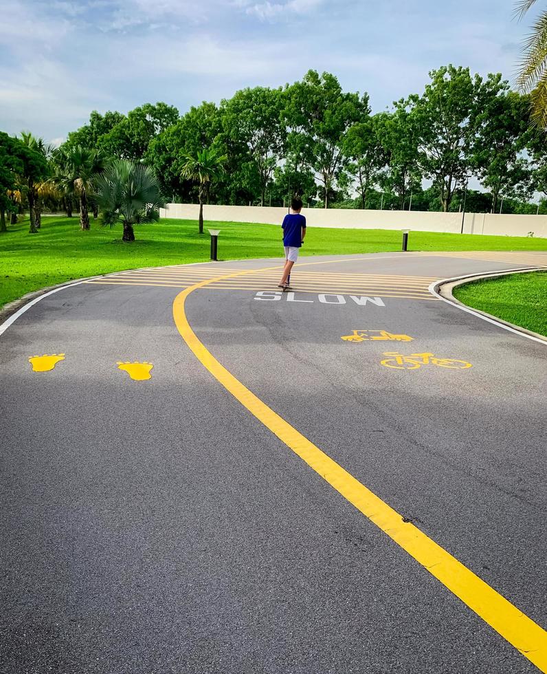 un niño sano se divierte montando scooter en el carril bici en el parque verde para hacer ejercicio. patinete infantil para el ocio en vacaciones. niño juega al aire libre. Patinete en carretera asfaltada con pasarela y carril bici. foto