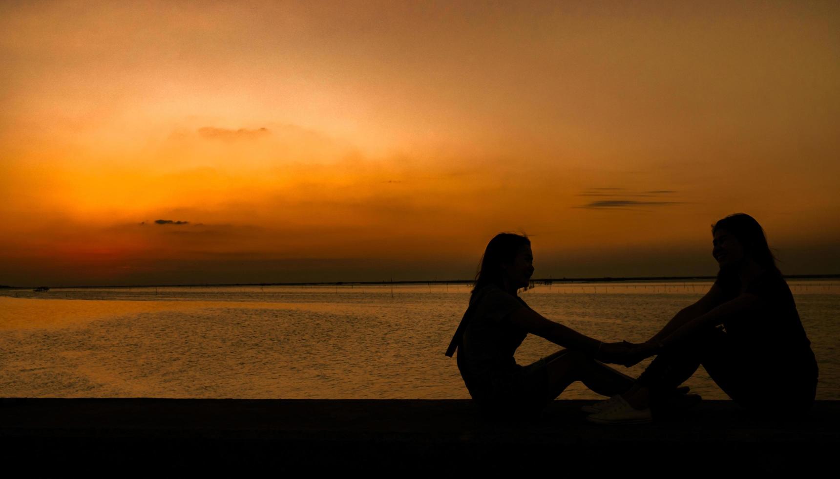 Silhouette of two pretty girl sit and holding hands and have fun together at the seaside at sunset with orange sky. Summer vacation and backpack travel concept. photo