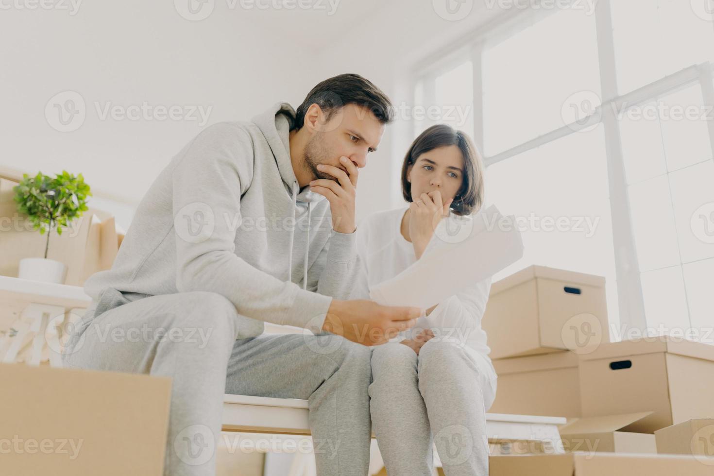Upset couple stressed by bad news, receive high taxes, looks attentively at papers, read paper letter, have to pay much for flat, move in other apartment, pack belongings in cardboard containers photo