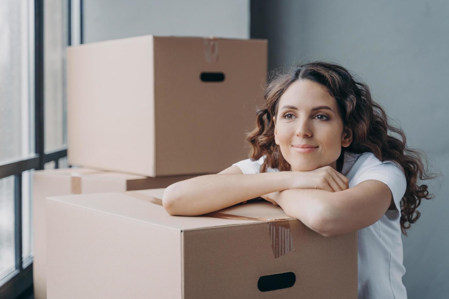 feliz mujer europea desempacando cosas inclinadas a cajas. chica soñando cerca de la ventana en un apartamento nuevo. foto