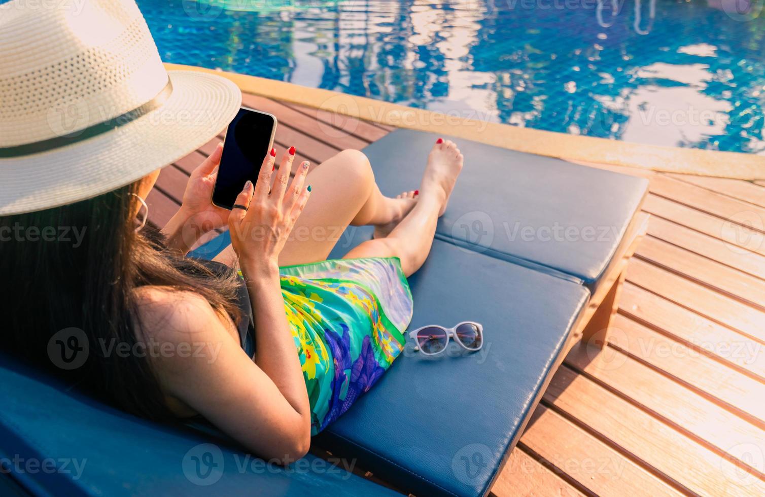 Asian woman with hat and swimsuit sit on sunbed at poolside and using smartphone on summer vacation by swimming pool. Girl with nail manicure and sunglasses. Summer vibes. Woman relaxing on holiday. photo