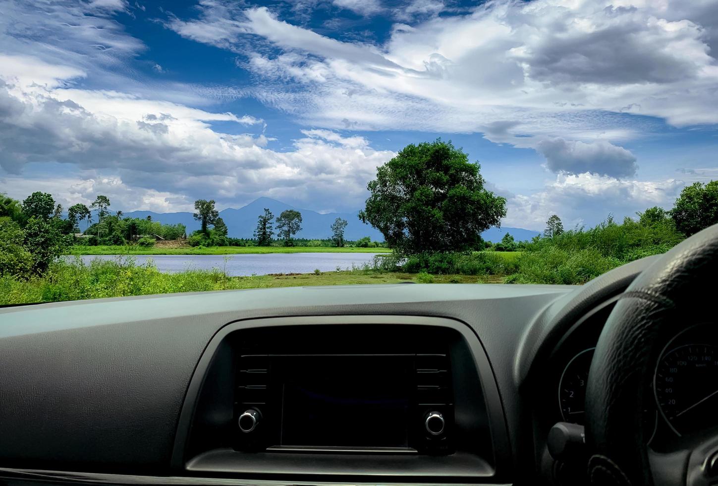 Beautiful landscape view from inside car. Steering wheel and dashboard of car interior. Road trip travel with scenic view of mountain, lake, and forest. Blue sky and white fluffy clouds. Vacation time photo