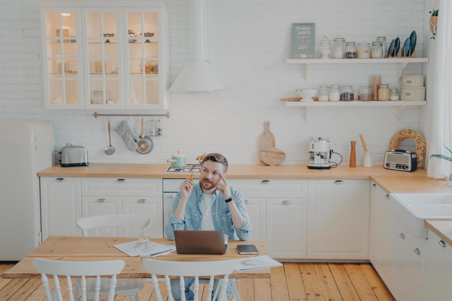 Young office worker working remotely from home with computer while sitting by kitchen table photo