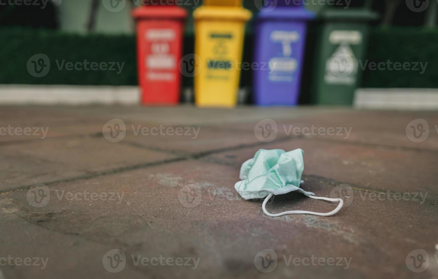 Used medical face mask discard on pavement floor on blurred recycle bin or trash. Medical waste disposal with unhygienic. Disposable earloop face mask waste garbage on floor. Green surgical face mask. photo