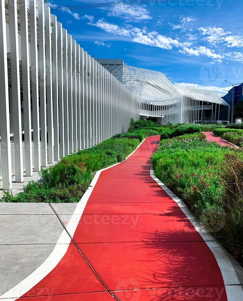 Empty red walkway for running in the garden near shopping mall. Running track in the park with green ornamental plant on sunny day. Public park for exercise and relaxation beside modern building. photo