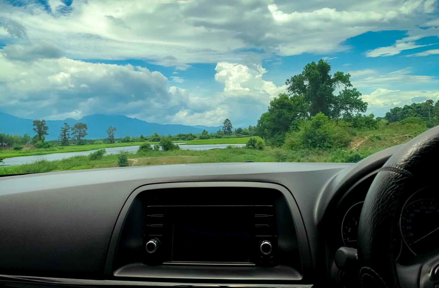 Beautiful landscape view from inside car. Steering wheel and dashboard of car interior. Road trip travel with scenic view of mountain, lake, and forest. Blue sky and white fluffy clouds. Vacation time photo