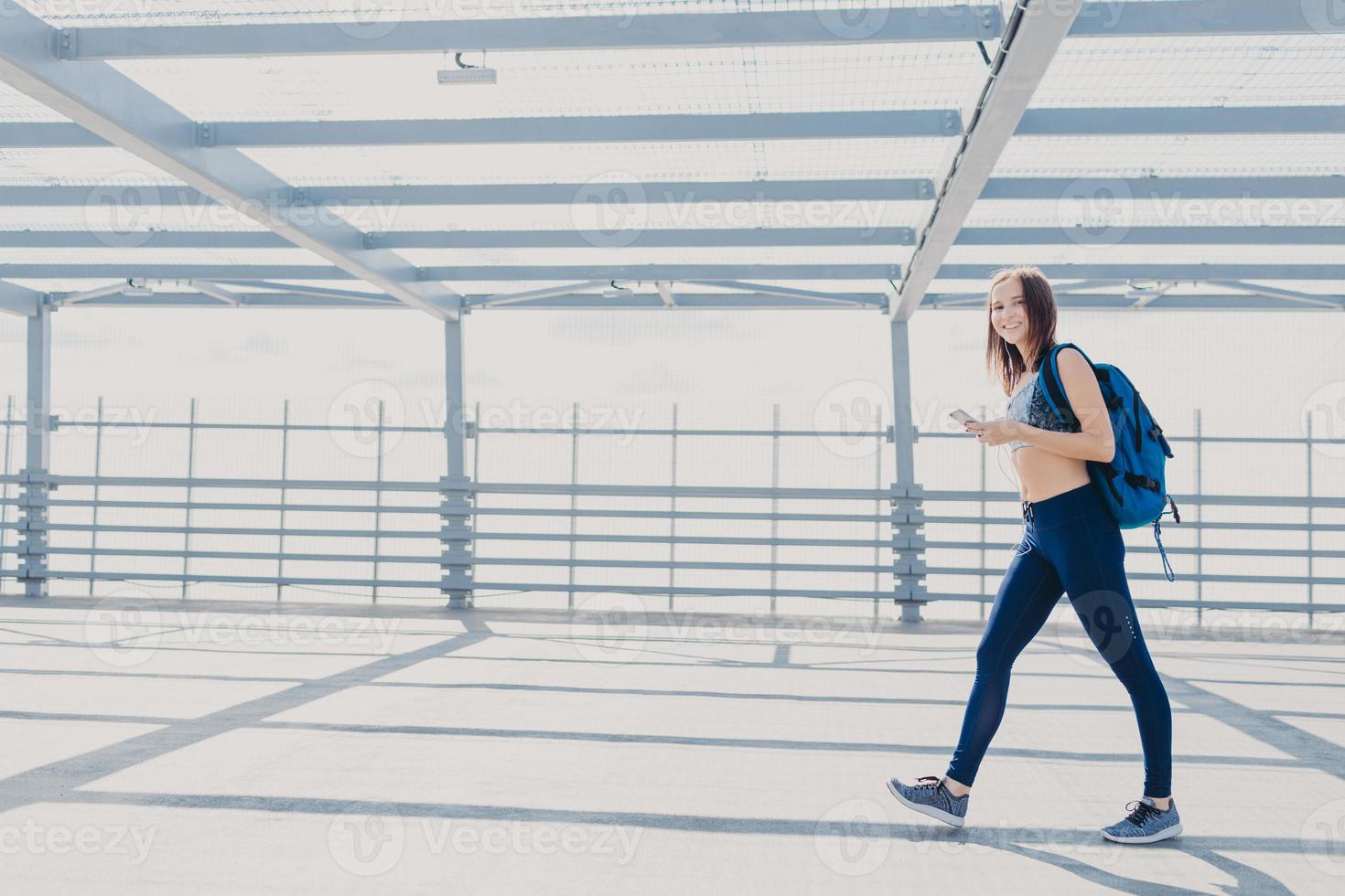 Active female fitness trainer dressed in sportsclothes, sneakers, carries rucksack, uses mobile phone for checking calories, listens music with earphones, uses free wifi connection. People and sport photo