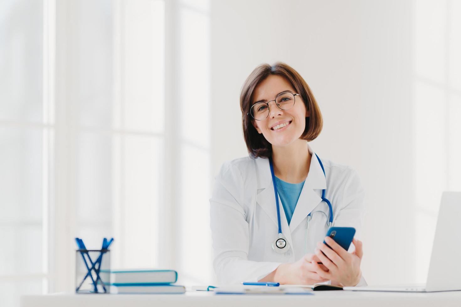 Horizontal view of smiling professional doctor works in clinic, poses at modern hospital office with electronic gadgets, sends text messages on cellphone being at work. Health care, technology concept photo