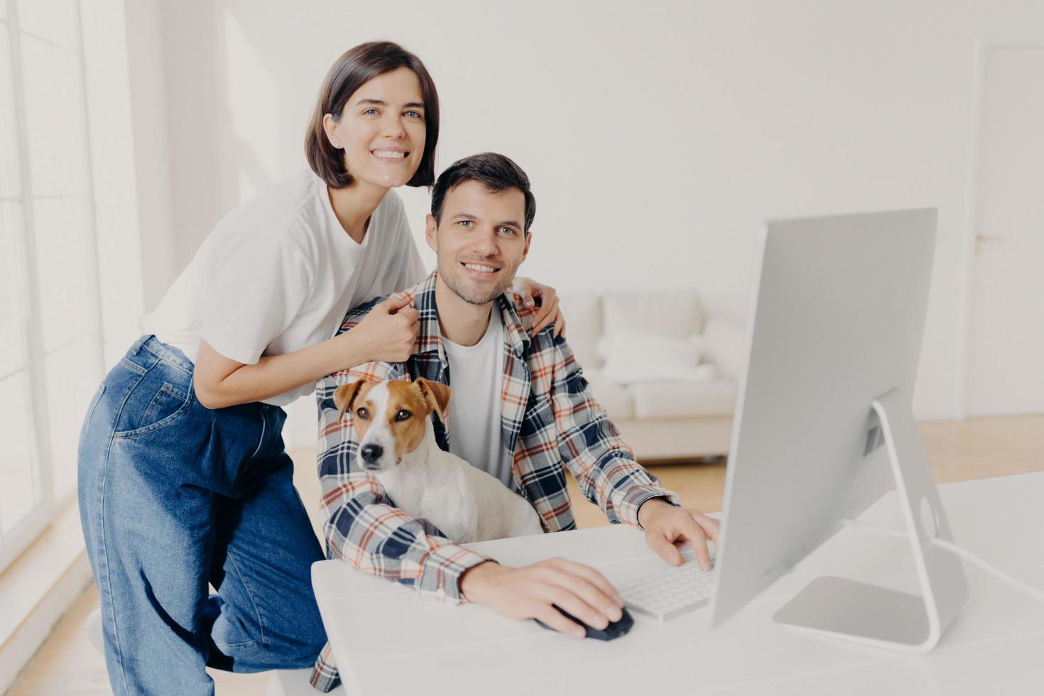 Image of happy brunette smiling woman leans at husbands shoulders who keyboards on computer, surf internet and search new apartment to buy on website, pose in spacious room with domestic dog photo