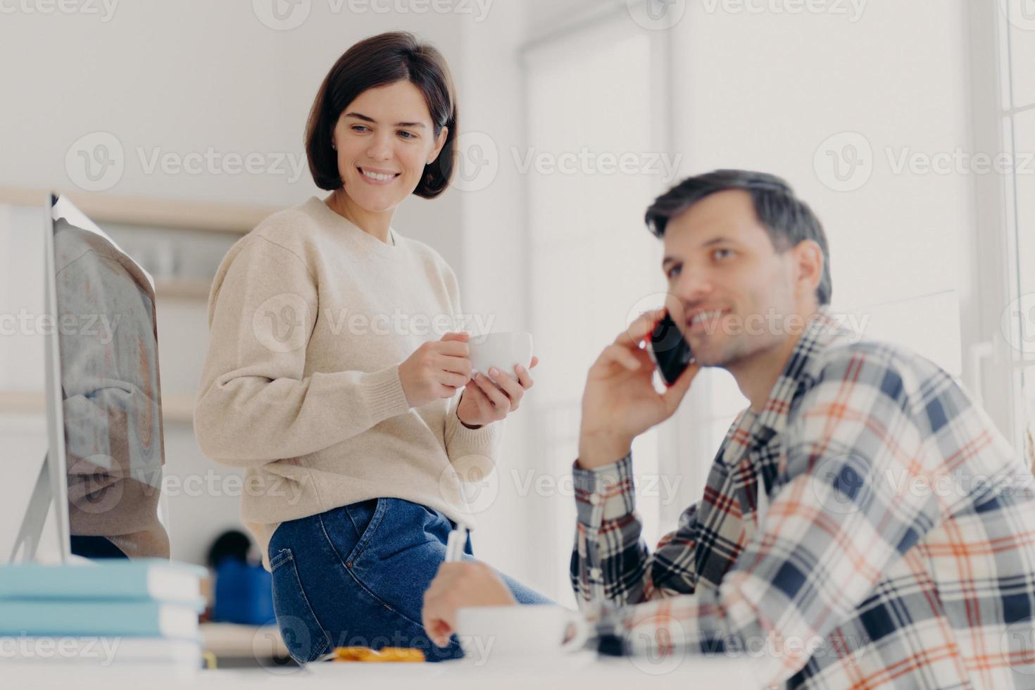 Young married couple study contract for new house, check documents for buying property, man discusses something with partner via cellphone, happy woman drinks tea and looks in computer monitor photo
