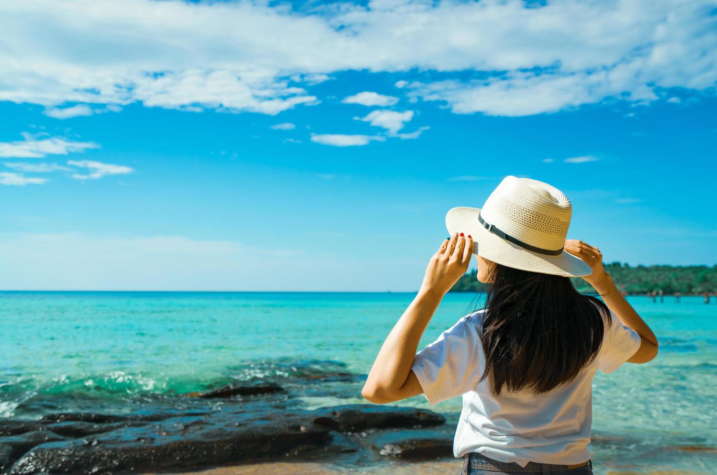 Happy young Asian woman in casual style fashion with straw hat stand at sea beach of resort in summer vacation . Relaxing and enjoying holiday at tropical paradise beach. Summer vibes. Summer travel. photo