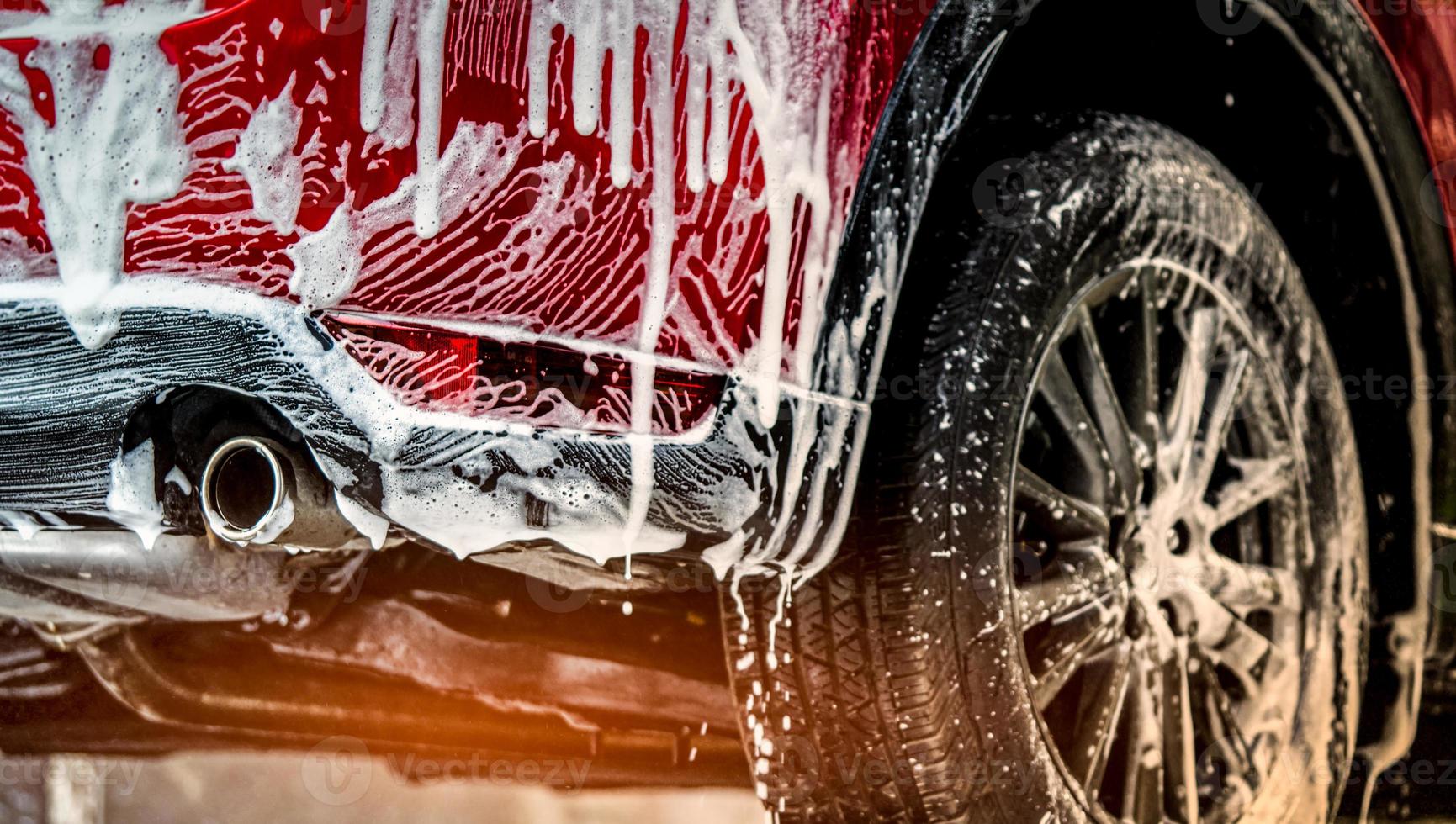 Red compact SUV car with sport and modern design washing with soap. Car covered with white foam. Car care service business concept. Car wash with foam before glass waxing and glass coating automobile. photo