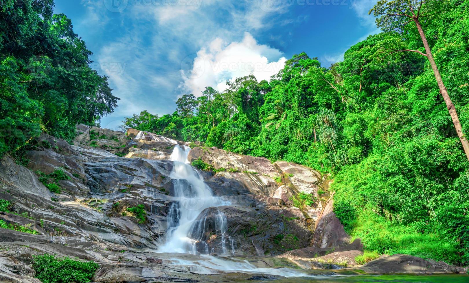 Beautiful waterfall at the mountain with blue sky and white cumulus clouds. Waterfall in tropical green tree forest. Waterfall is flowing in jungle. Nature abstract background. Granite rock mountain. photo