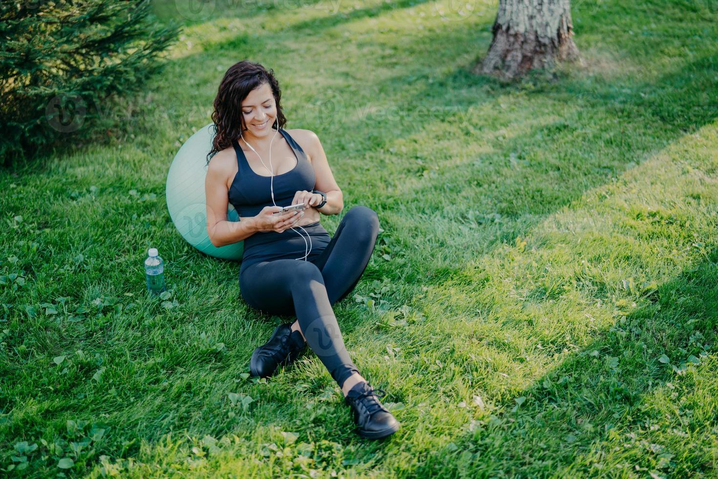 Relaxed sportswoman has rest after doing sport exercises with fitball, uses smartphone, listens favorite music in earphones from playlist, sits on green grass, bottle of fresh water near. Aerobics photo